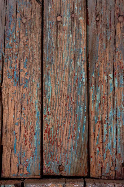 Kırsal ahşap kalaslar kırmızımsı kahverengi, deniz mavisi ve solmuş açık turuncu tonlarında kabuk soyan boyaları sergiler. Çiviler görünür, gün ışığında yaşlanmış, dokulu yüzeyi vurgular..