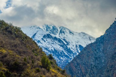 Kuru bitki örtüsüyle kaplı dik yamaçlı karlı dağların dramatik manzarası. Bulutlu gökyüzü bu dağlık bölgede sakin ve soğuk bir atmosfer yaratır..
