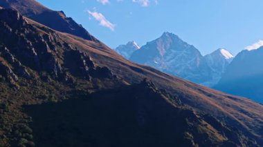 Kırgızistan 'daki dağ manzarasının hava görüntüleri. Dik, kayalık bir yamaç ön plana hükmediyor, dağların kıyısındaki bir vadiye iniyor. Uzakta karla kaplı zirveler dağınık bulutlu açık mavi bir gökyüzüne karşı.