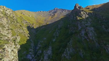 Drone görüntüleri Kırgızistan 'daki dramatik bir dağ vadisini, kayalık kayalıkları, yeşil yamaçları ve açık mavi gökyüzünün altında uzak bir zirveyi yakalar. Kamera soldan sağa hareket ediyor, parallax boyunca derinliği gösteriyor.