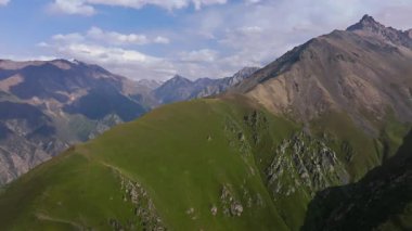 Kırgızistan 'ın engebeli dağ manzarasını gösteren hava aracı görüntüleri. Parçalı bulutlu gökyüzünün altındaki yeşil vadiler ve kayalık tepeler bölgenin doğal güzelliğini gözler önüne seriyor..