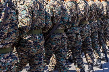 Çok sayıda asker, askeri geçit sırasında disiplin ve düzen sergileyerek düzen içinde yürür. Kamuflaj üniformaları ve tüfekler açık havada ciddi ve resmi bir atmosferi yansıtıyor. Arka görünüm.