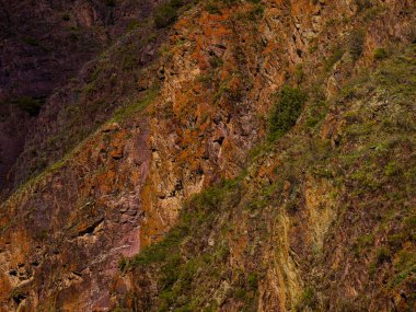 Çeşitli bitki örtüsüyle süslenmiş engebeli kayalık oluşumlar dağlık bir bölgede dramatik bir manzara yaratır. Doğal dokular ve renkler açık hava manzarasının vahşi güzelliğini vurgular..