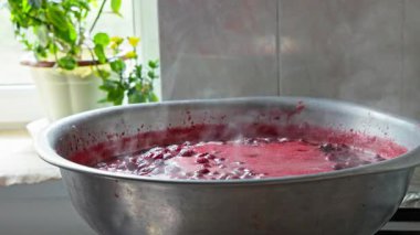 Large metal pot simmers with boiling red strawberry confiture in warm kitchen. Steam rises near window, creating a cozy domestic atmosphere.