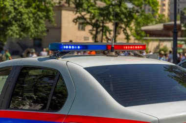 White police sedan with red and blue emergency lights illuminated. Blurred figures of people in background create an alert and suspenseful atmosphere in an urban environment.