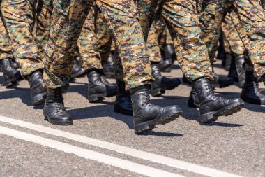 Soldiers dressed in camouflage trousers and black boots march in formation on an asphalt road at sunny summer day.