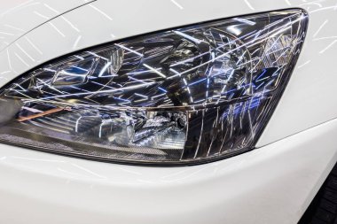 Shiny car headlight captures reflections of bright lights in a modern garage. Clean lines and sleek design enhance the contemporary feel of the indoor setting.