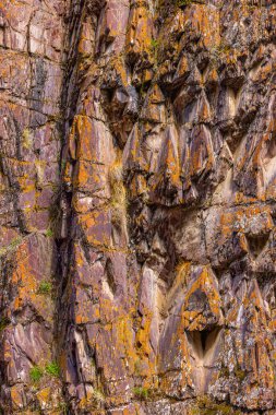 Engebeli kaya oluşumu turuncu liken aksanlı canlı mor ve kahverengi renkler sergiliyor. Benzersiz dokular ve şekiller, bu doğal açık hava ortamının vahşi güzelliğini vurgular..