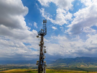 Haberleşme kulesi engin kırsal alanlarda, tepeler ve dağlarla birlikte yüksekte duruyor. Uydu çanakları ve antenler parçalı bulutlu gökyüzüne karşı teknolojiyi vurguluyor.