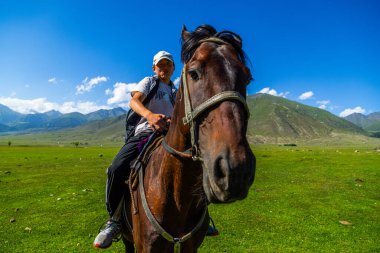 Genç adam manzaralı dağlık arazide kahverengi ata biniyor. Parlak gün ışığında, koyunların otladığı huzurlu bir atmosfer. Semenovskoye Boğazı, Kırgızistan - 17 Temmuz 2024