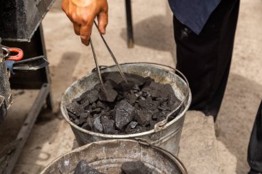Odaklanmış adam açık hava atölyesinde kömür toplamak için maşa kullanıyor. Kasıtlı eylem, kırsal çalışma ortamını ve çalışkan ruh halini yansıtır..