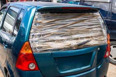 Arka camı hasarlı, temiz bantla kaplı bir araba. Gündüz sahnesi geçici bir onarım çözümünü ya da yetersizliği ve tamir masraflarını karşılayamamayı yansıtıyor.