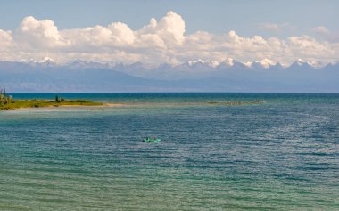 İki insan, sakin bir Issyk-Kul gölünde küçük bir teknede kürek çekerken huzurlu bir anın tadını çıkarıyor. Karla kaplı dağlar ve uzak kıyı şeridi sakin bir zemin oluşturur.