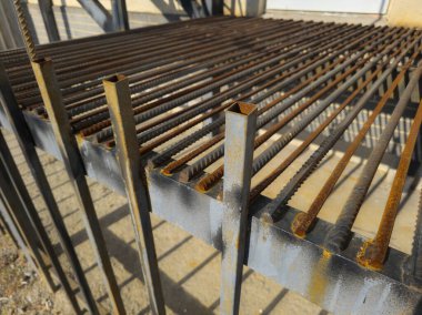 Paslı çelik ızgara, açık havada aşınma ve yırtık gösteriyor. Güneş ışığı paslanmış çubukları ve destekleri vurguluyor ve kırsal bir endüstriyel atmosfer yaratıyor..