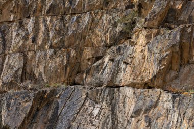 Engebeli kaya oluşumu, küçük ve dayanıklı bitkilerle çevrili dağlık bir bölgede gururla duruyor. Topraksı tonlar ve dokular gündüz vakti çorak ama çarpıcı bir doğal ortam yaratır..