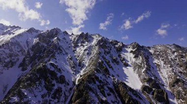 Karla kaplı yamaçları ve açık mavi gökyüzünün altında kayalık arazisi olan görkemli bir dağ sırası. Soldan gelen hareket ile dron görünümü.