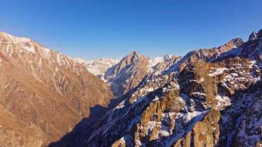 Yüksek rakımlı dağ sıraları açık mavi gökyüzüne karşı karla kaplı zirveleri sergiliyor. Yan hareket ile dron görünümü.