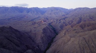 İnsansız hava aracı nehir yatağı boyunca sarmal yeşil bitki örtüsüyle dik kayalık vadide süzülüyor. Çorak kırmızımsı kahverengi yamaçlar her iki tarafta keskin bir şekilde yükseliyor, kırgızistan 'da engin dağlık arazi alacakaranlıkta..