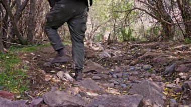 Yetişkin bir adam huzurlu bir ormanda kayalık bir yolda kararlı bir şekilde yürür. Yürüyüş botları ve pantolonları içinde maceraperest bir ruhu simgeliyor..