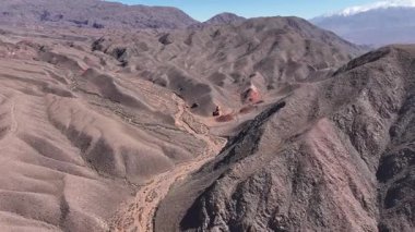 Havadan bakıldığında kayalık kırmızı oluşumlar ve kuru bir nehir yatağının yer aldığı engin kurak bir manzara görülür. Arka planda görkemli dağlar yükselir ve sakin bir atmosfer yaratır..