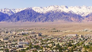 Kırgızistan 'daki görkemli dağların eteklerine yerleşmiş uçsuz bucaksız şehir manzarasını görüntüler. Gündüz sahnesi, sakin doğanın kentsel gelişimle yan yana olduğunu ortaya koyuyor..