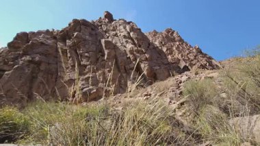 Kayalık dağ yüzü kurak manzaraya karşı dimdik ayakta duruyor. Kuru otlar ve dağınık çalılar gün ışığında engebeli ve ıssız araziyi güçlendirir..