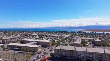 Gökyüzü manzarası, heybetli dağların çerçevelediği sakin göl kenarındaki geniş şehir manzarasını yakalar. Açık mavi gökyüzü huzurlu gündüz atmosferini artırır.