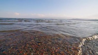 Serene Issyk-Kul göl manzarasında parlak gün ışığında pürüzsüz çakıl taşları ve temiz su yer alıyor. Huzurlu doğa ayarı gevşemeyi ve yansımayı davet eder.