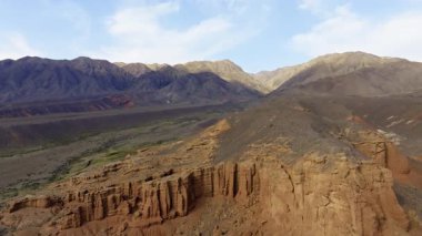 Gökyüzü görüntüsü Kırgızistan 'daki engin, engebeli dağları ve kumtaşı kaya oluşumlarını yakalar. Huzurlu vadi manzarası seyrek bitki örtüsü ve bulutlu berrak bir gökyüzü..