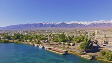 Hava manzaralı geniş bir mavna, Issyk-Kul Gölü 'ndeki Balykchy kentindeki sanayi binaları tarafından rıhtıma yanaşmış çok sayıda tekne görülüyor. Ağaçlar ve arka planda karla kaplı dağlarla sakin bir atmosfer..