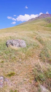 Serene sahnesinde rüzgarda sallanan uzun otlarla çevrili toprak bir patika boyunca büyük bir kaya var. Dağlar yükseliyor mavi gökyüzünün altında dağınık bulutlarla.