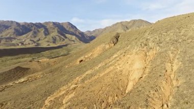 Kırgızistan 'da hava manzarası, açık gökyüzü altında erozyon desenleri, kuru bitki örtüsü ve kayalık tepeler gösteren uçsuz bucaksız kurak bir manzara yakalar. Dolly geri kamera hareketi.