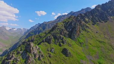 Uçsuz bucaksız dağların uçsuz bucaksız görüntüsü berrak mavi gökyüzüne karşı görkemli bir şekilde yükseliyor. Barışçıl manzarada kayalık arazi ve açık havada yemyeşil çimenler bulunur. Kamyon sol kamera hareketine.