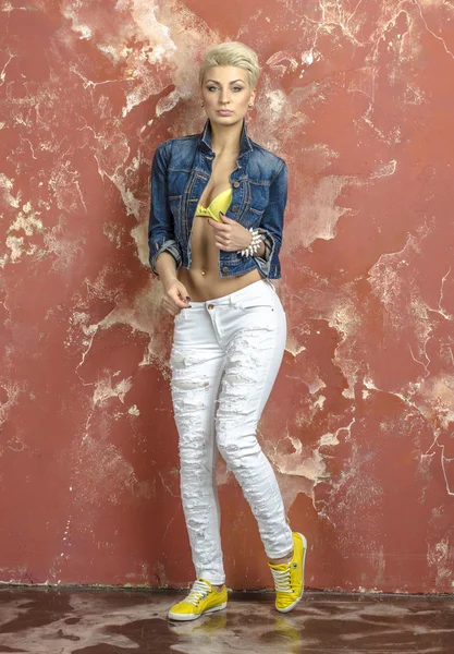 Beautiful blonde with short hair in a yellow lingerie and jeans takes denim jacket — Stock Photo, Image