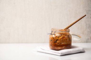 Geleneksel mayalanmış Kore kimchi lahanasıyla dolu cam kavanoz beyaz bir tekstil peçetesine ahşap çubuklarla yerleştirilmiş. Parlak, minimalist yemek fotoğrafçılığı ve temiz beyaz desenli duvar arkaplanı. Yüksek açılı görünüm, boşluğu kopyala