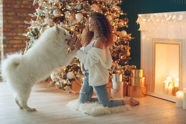 Hermosa mujer con abrazos de perro, sobre el fondo de una chimenea y un árbol de Navidad con velas. —  Fotos de Stock