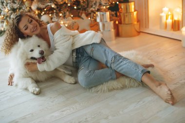 Güzel bir kadın, süslenmiş bir odada mumlarla, bir Noel ağacı şöminesinin arka planında, beyaz bir battaniyede köpek kucaklamalarıyla oturuyor. Mutlu yıllar ve mutlu Noeller..