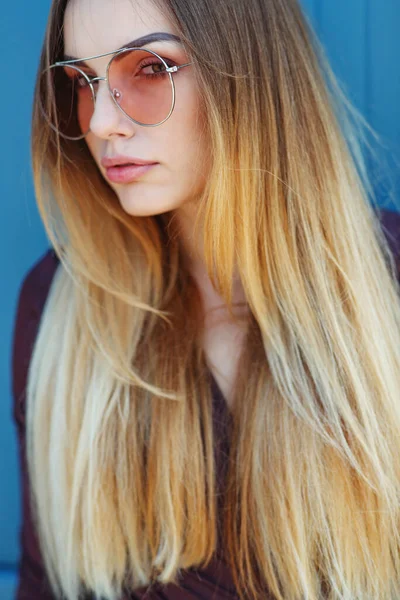 Nahaufnahme einer eleganten jungen Frau mit Sonnenbrille, die vor blauem Hintergrund lächelt. Schöne weibliche Modell mit Kopierraum. — Stockfoto