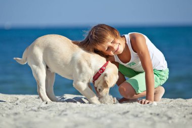 Mutlu çocuk mavi gökyüzüne karşı deniz kıyısında köpeğiyle oynuyor. En iyi arkadaşlar tatilde eğlenir, denize karşı kumsalda oynarlar..