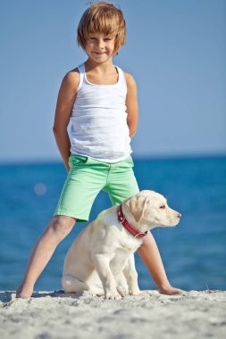 Mutlu çocuk mavi gökyüzüne karşı deniz kıyısında köpeğiyle oynuyor. En iyi arkadaşlar tatilde eğlenir, denize karşı kumsalda oynarlar..