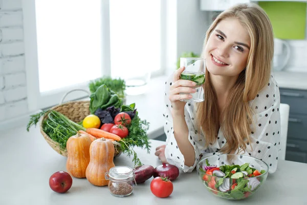 Giovane donna che beve acqua vicino al tavolo con frutta e verdura in cucina. Cibo sano, bevande, dieta, disintossicazione e concetto di persone. — Foto Stock
