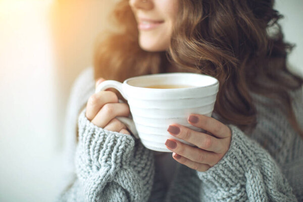 Close-up of female hands with a mug of drink. Beautiful girl in a gray sweater is holding a cup of tea or coffee in the morning sunlight. Mug for your design. 