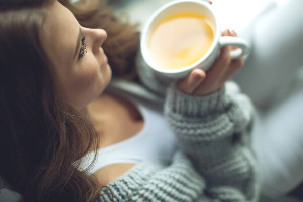 Close-up of female hands with a mug of drink. Beautiful girl in a gray sweater is holding a cup of tea or coffee in the morning sunlight. Mug for your design. 