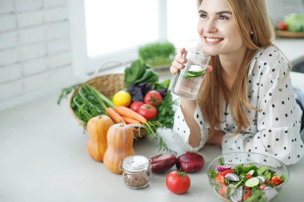 Ung kvinna dricksvatten nära bordet med frukt och grönsaker i köket. — Stockfoto