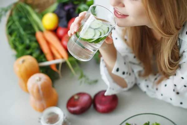 Ung kvinna dricksvatten nära bordet med frukt och grönsaker i köket. — Stockfoto