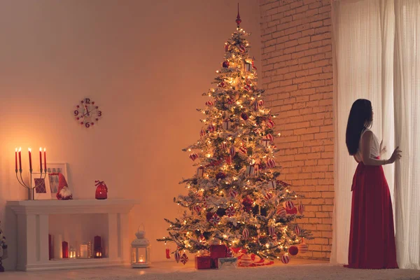 Mujer en un hermoso vestido, noche cálida y acogedora en el diseño de interiores de Navidad, árbol de Navidad decorado con luces regalos juguetes, velas, linternas, iluminación de guirnaldas en la sala de la chimenea. — Foto de Stock