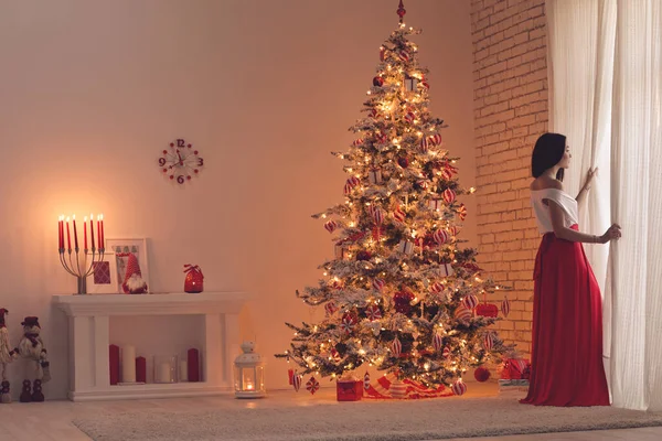 Γυναίκα σε ένα όμορφο φόρεμα, ζεστό και ζεστό βράδυ στο χριστουγεννιάτικο εσωτερικό σχεδιασμό, χριστουγεννιάτικο δέντρο διακοσμημένα με φώτα δώρα παιχνίδια, κεριά, φανάρια, φωτισμός γιρλάντα στην αίθουσα τζάκι. — Φωτογραφία Αρχείου