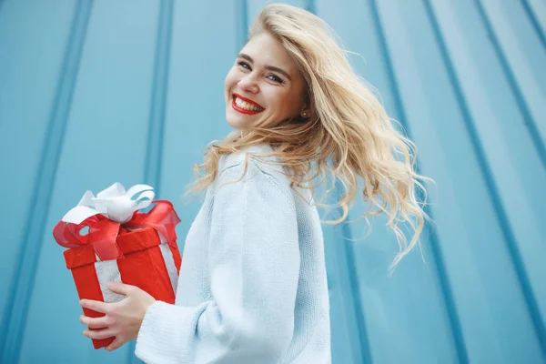 Heureuse femme blonde en tenant boîte cadeau et en regardant la caméra tout en appréciant sur fond bleu. — Photo