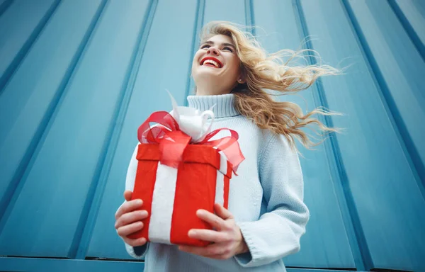 Boldog szőke nő kezében ajándék doboz és nézi a kamera, miközben élvezi a kék háttér. — Stock Fotó