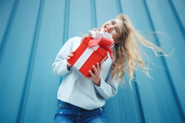 Heureuse femme blonde en tenant boîte cadeau et en regardant la caméra tout en appréciant sur fond bleu. — Photo